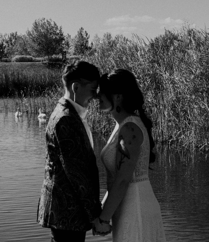 fotografia en blanco y negro de una pareja de chicas el dia de su boda agarrandose las manos y juntando las frentes en un lago con cisnes